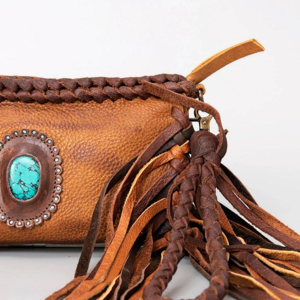 Close-up of a western-style leather clutch with braided details, fringes, and a turquoise stone centerpiece, showcasing fine craftsmanship.