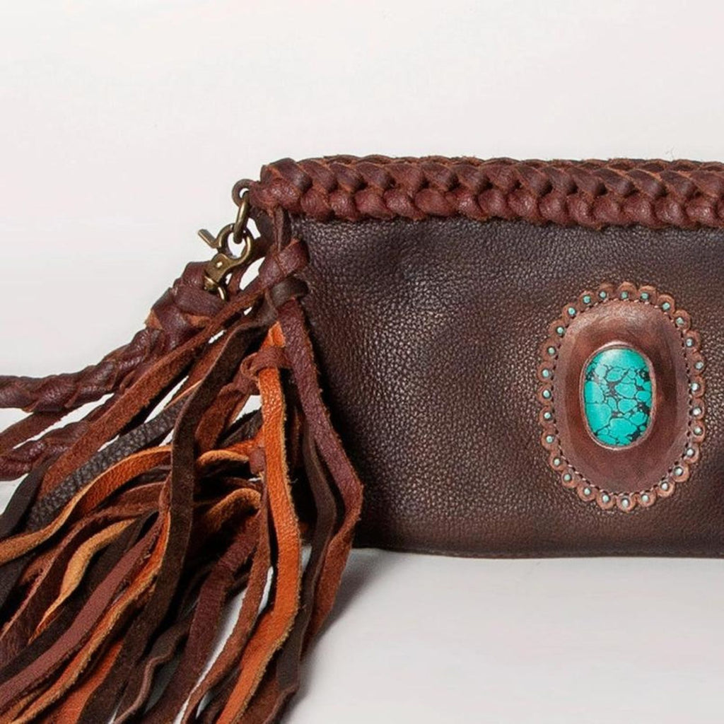 Close-up of a dark brown western-style leather clutch with braided details, fringes, and a turquoise stone centerpiece, highlighting its southwestern charm.