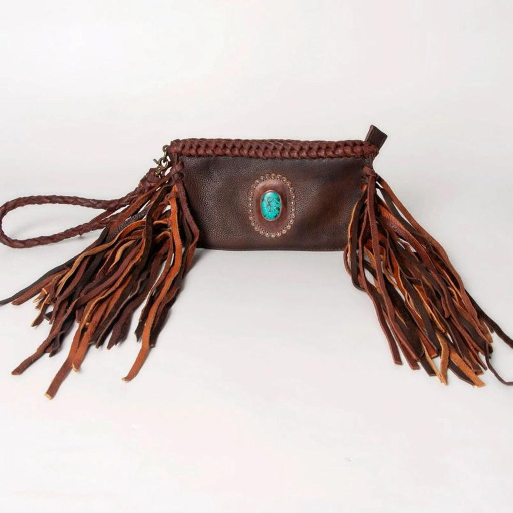 A dark brown western-style leather clutch with braided details, long fringes, and a turquoise stone centerpiece, evoking a southwestern aesthetic.