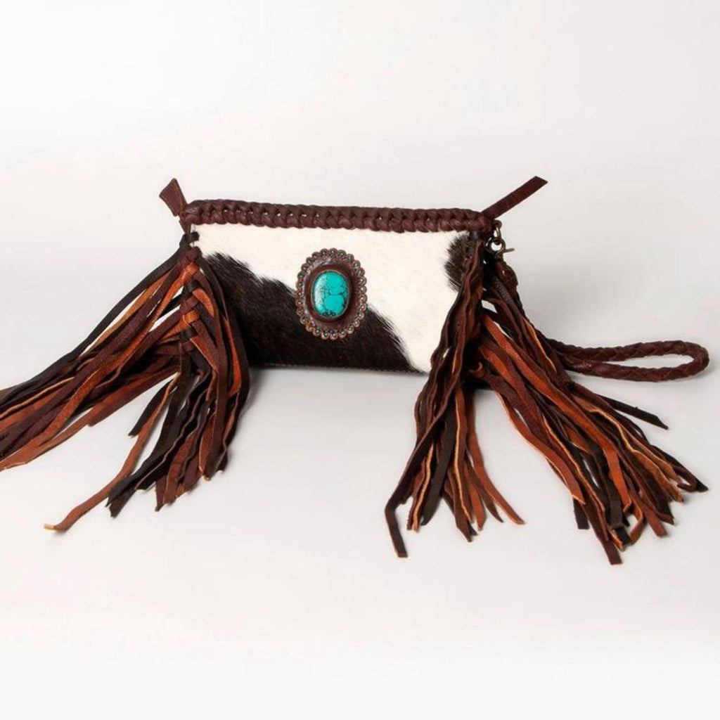 A handcrafted western-style leather clutch featuring hair-on leather with brown and white tones, long fringes, and a turquoise stone centerpiece.