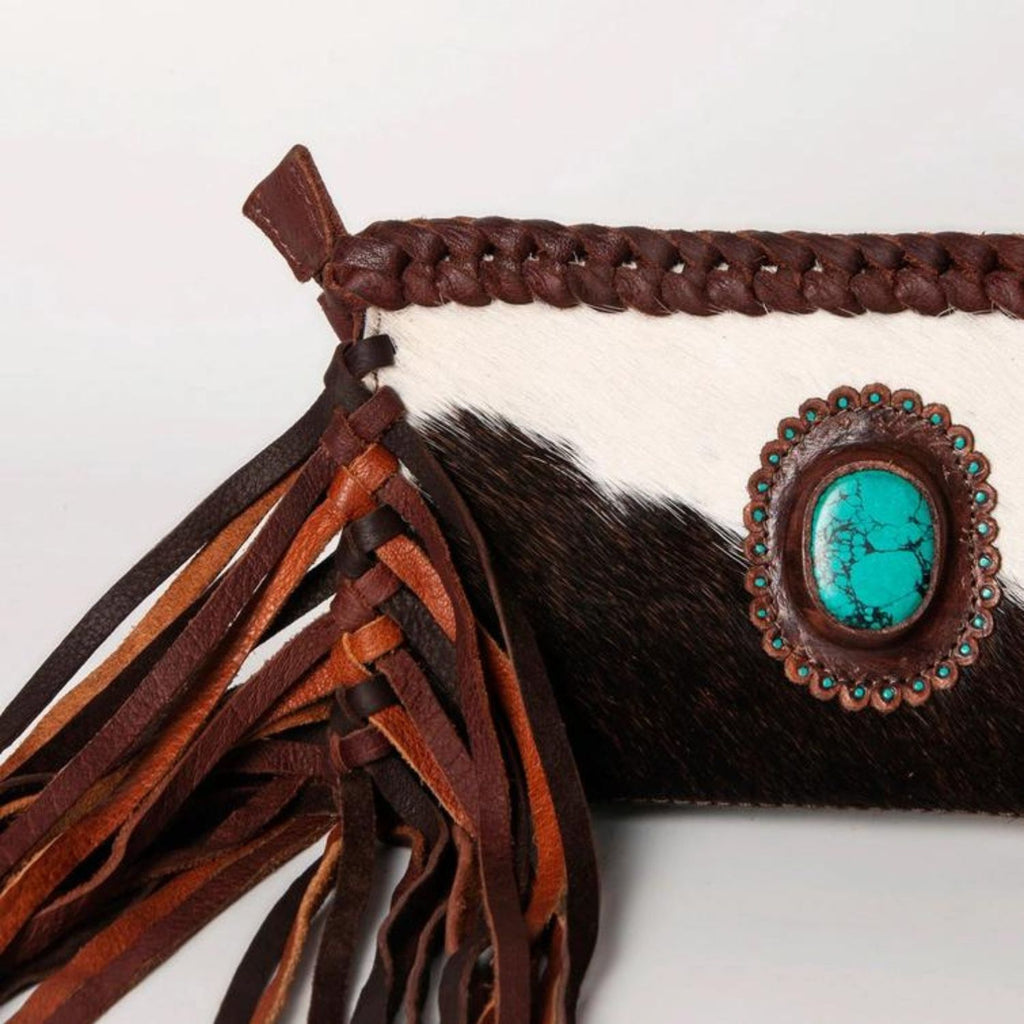 Close-up of a handcrafted hair-on leather clutch featuring braided details, fringes, and a turquoise stone centerpiece.