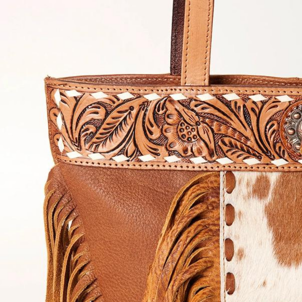 Close-up of hand-tooled floral detailing and hair-on cowhide leather on a Western-style tote bag with fringes.