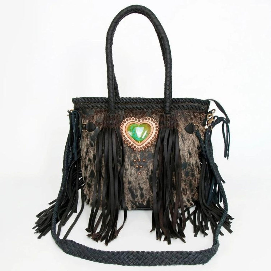 A black hair-on leather tote featuring braided leather handles, fringes, and a heart-shaped green stone centerpiece, designed by American Darling.