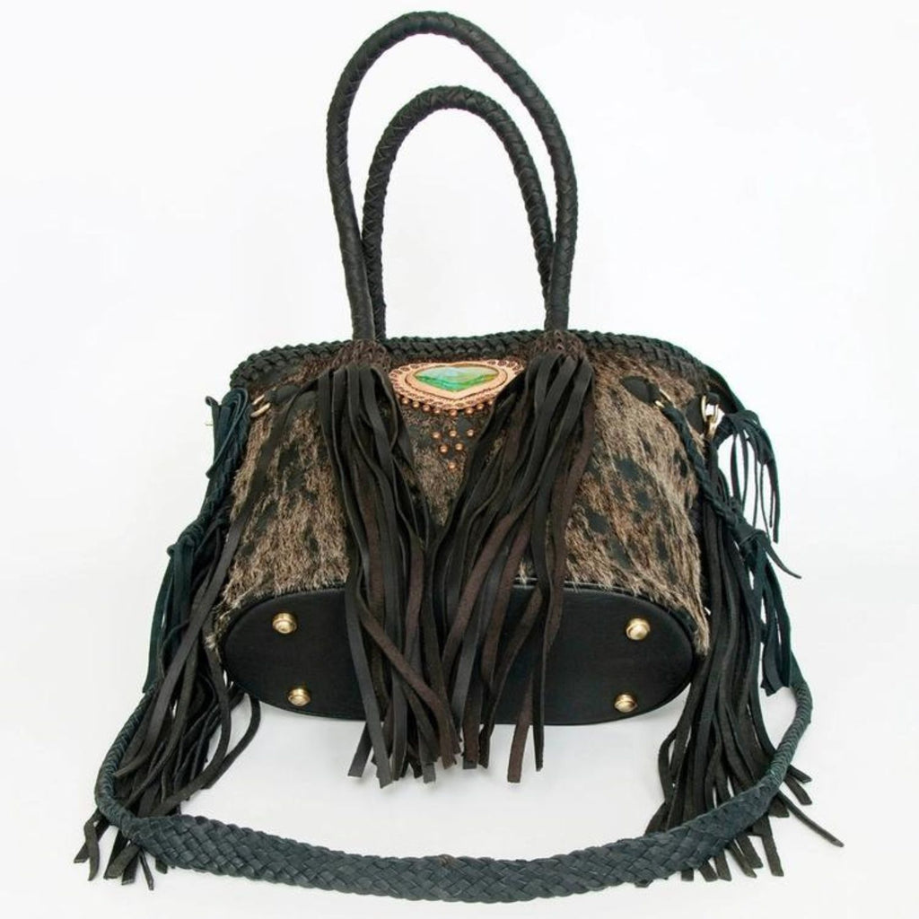 The bottom view of a black hair-on leather tote featuring braided leather handles, fringes, and protective metal feet, designed by American Darling.