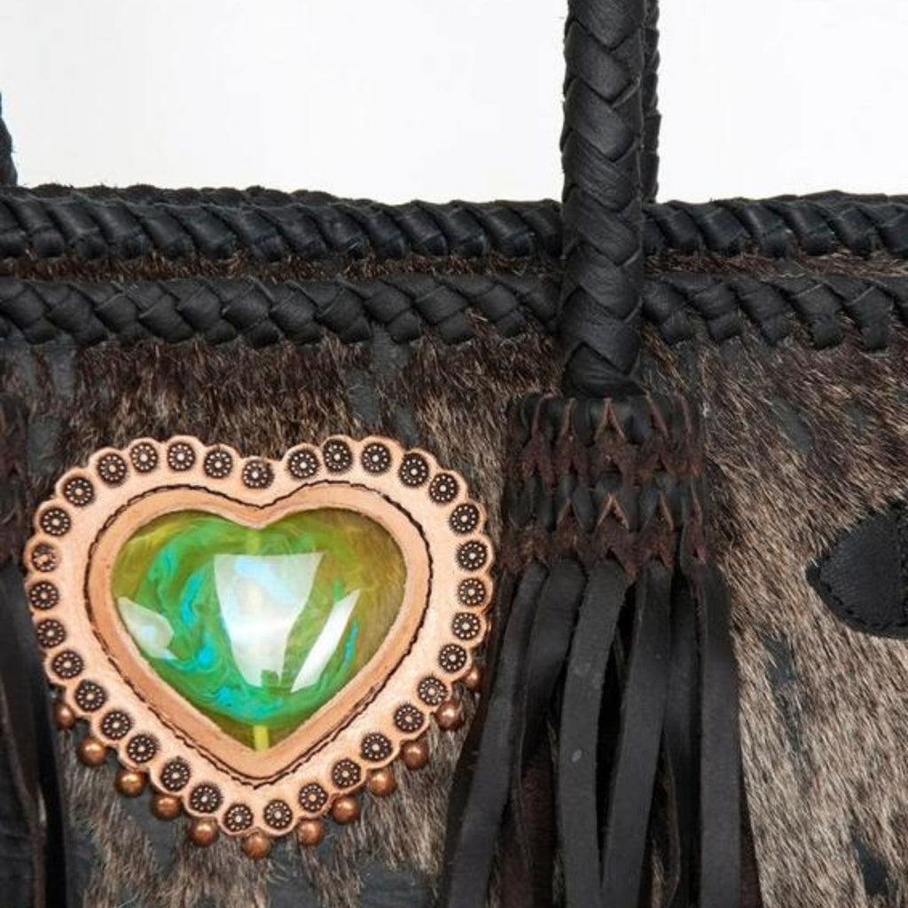 Close-up of the heart-shaped green stone embellishment on a black hair-on leather tote with braided leather handles and fringes, designed by American Darling.