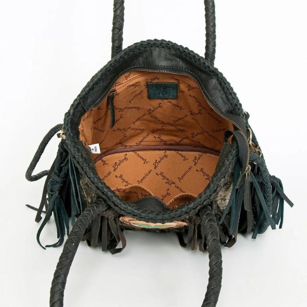 The interior view of a black hair-on leather tote featuring branded fabric lining, a zippered pocket, and braided handles, designed by American Darling.