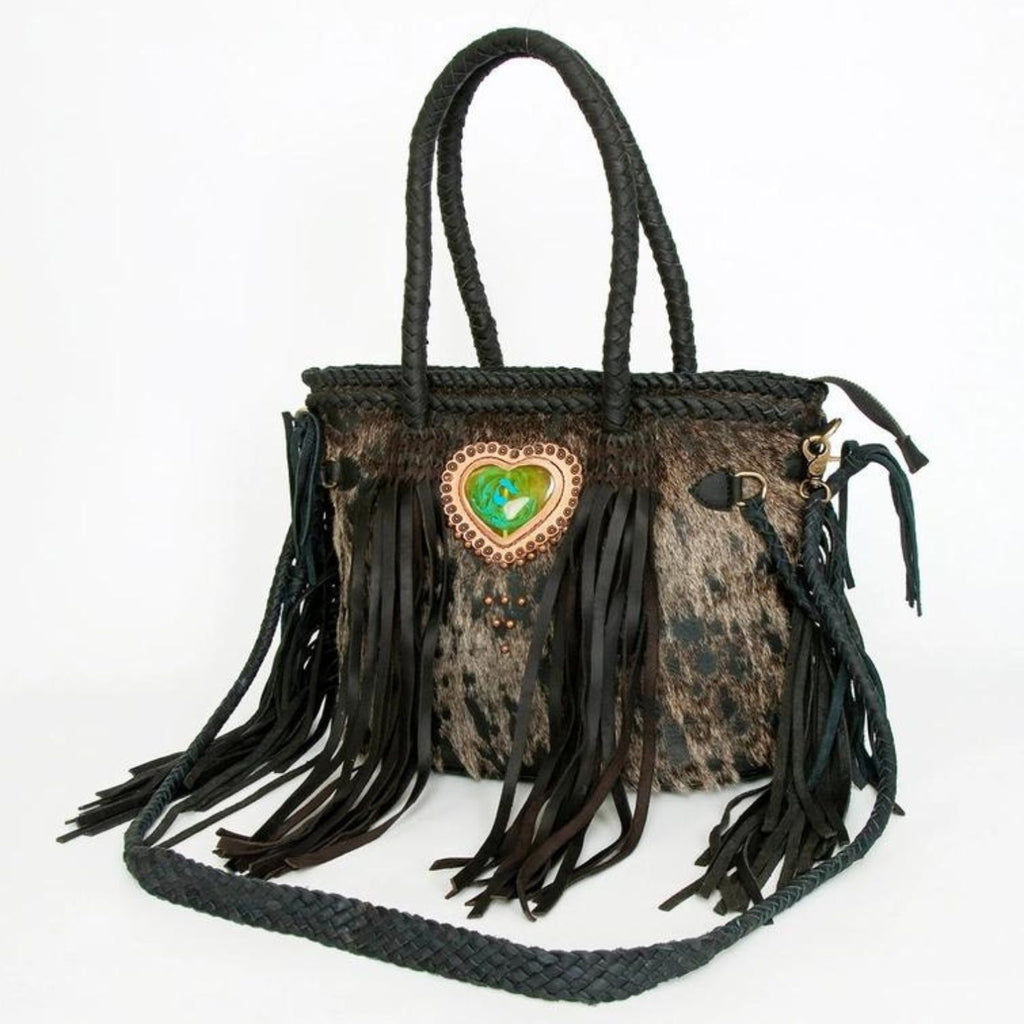 A handcrafted hair-on leather tote featuring black leather braiding, fringes, and a heart-shaped green stone centerpiece, designed by American Darling.