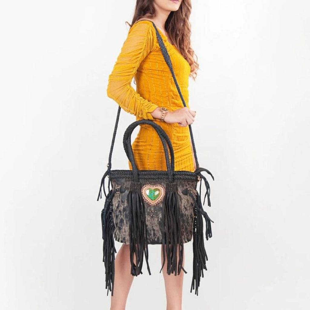 A model wearing a yellow dress holding the Heartland Hair-On Braided Leather Tote with black leather fringes and a heart-shaped green stone centerpiece.