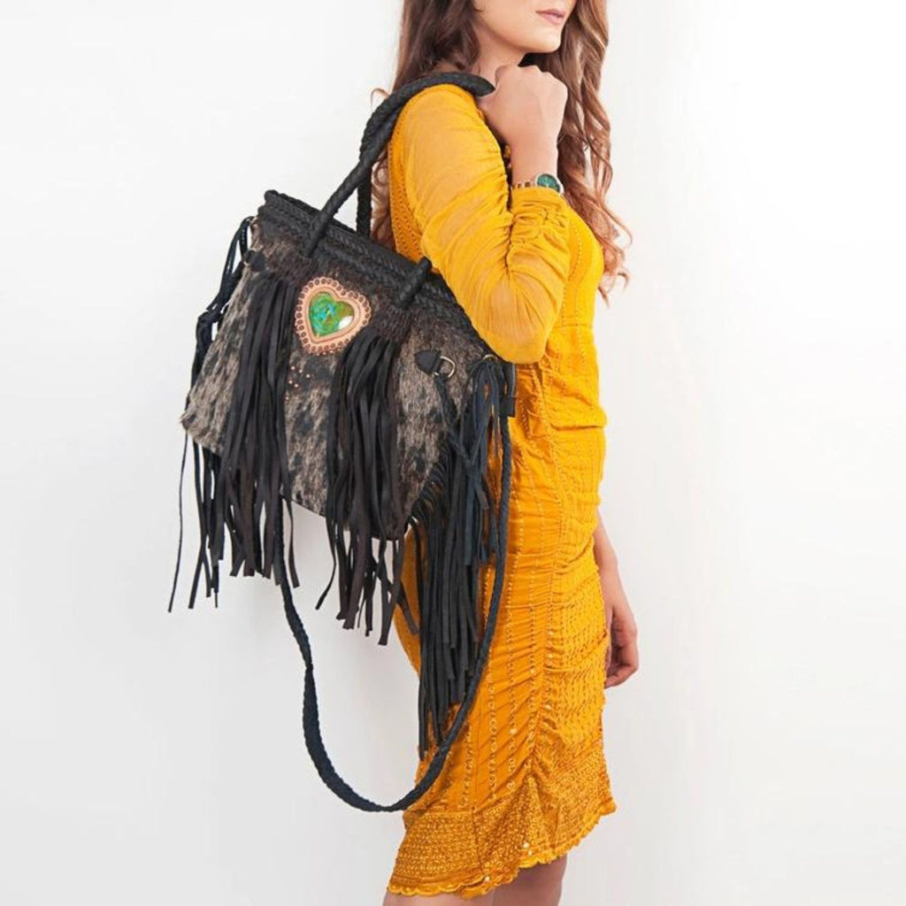 A model wearing a yellow dress carrying the Heartland Hair-On Braided Leather Tote with black leather fringes and a heart-shaped green stone.