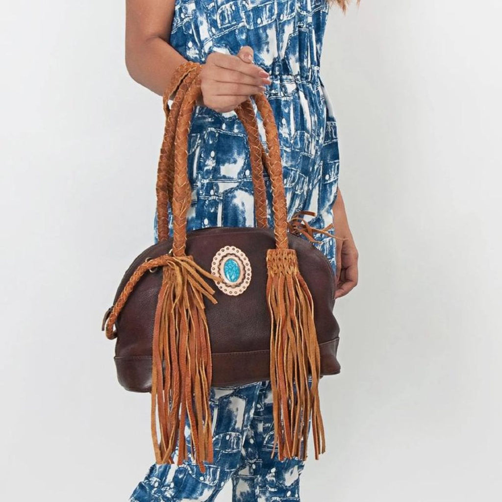 Model holding the Turquoise Trail Fringed Leather Tote by its braided handles, showcasing the leather fringe and turquoise stone centerpiece in a stylish Western design.